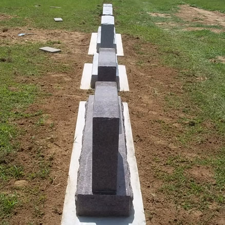 A row of newly installed granite headstones on concrete foundation pads in a cemetery, neatly aligned on a grassy and dirt-covered field.